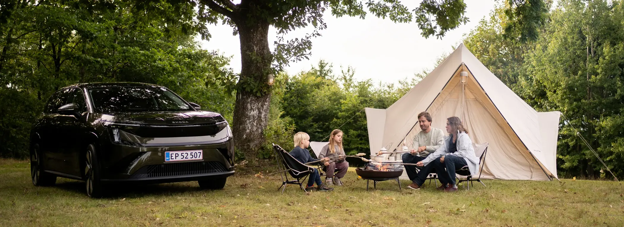 En sort Škoda SUV holder parkeret ved siden af en familie, der hygger sig omkring et bål foran et stort lyst telt i naturen.