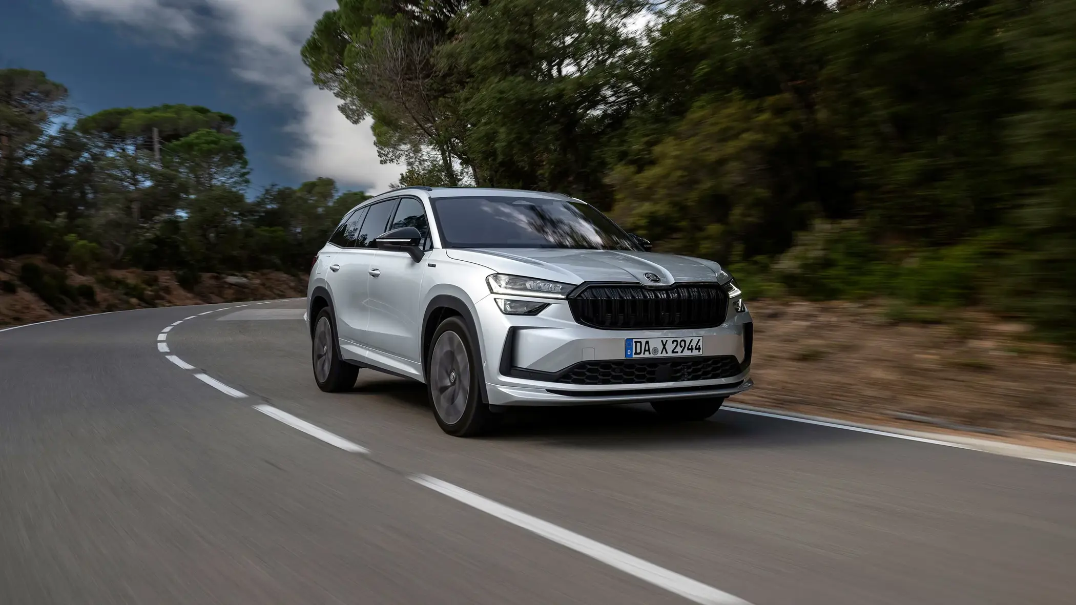 En sølvfarvet Škoda Kodiaq Sportline SUV kører hurtigt gennem et sving på en landevej omgivet af grøn skov.