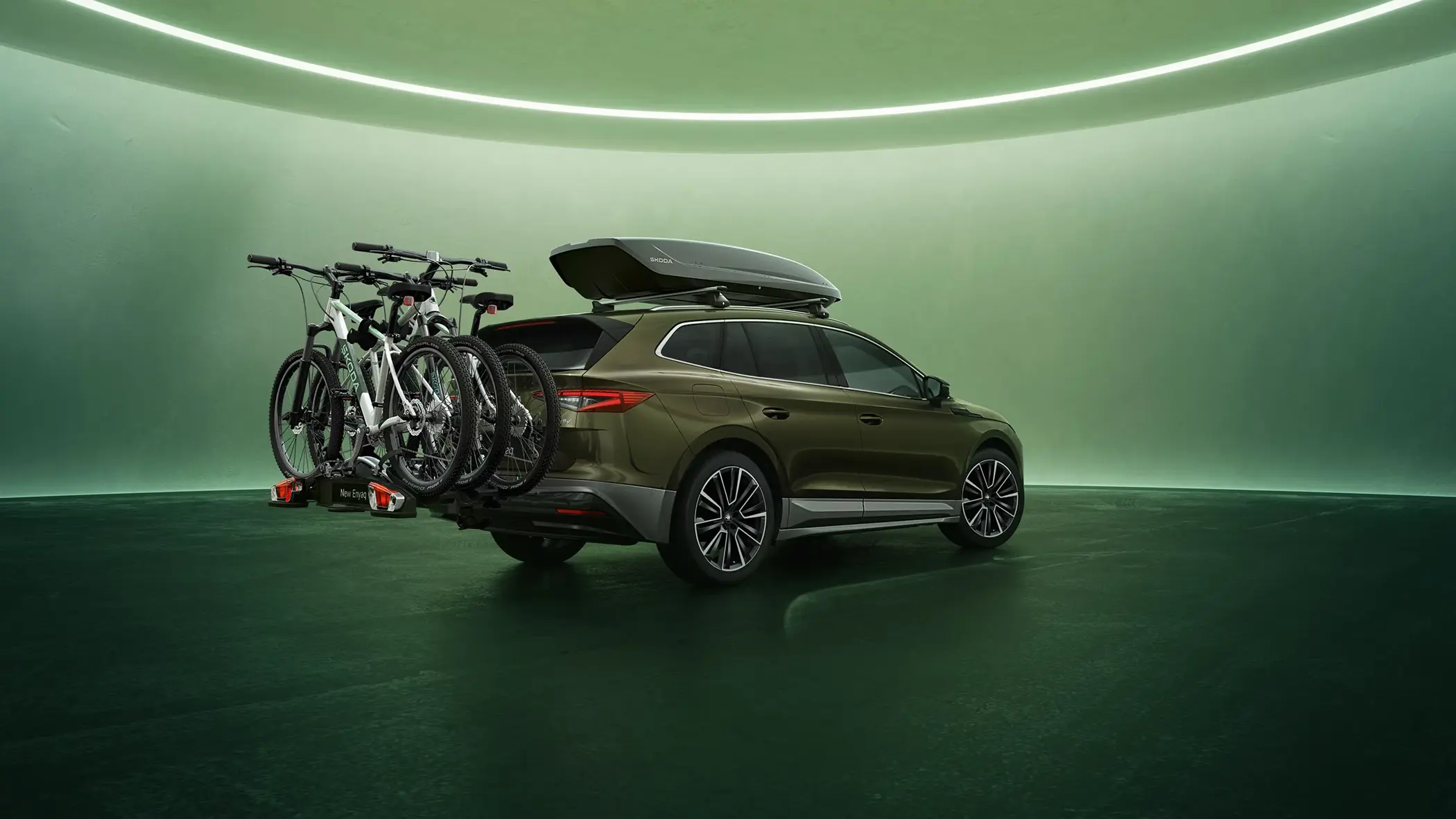 En Škoda Enyaq med tagboks og cykelholder med to cykler står i et grønt, futuristisk oplyst rum.